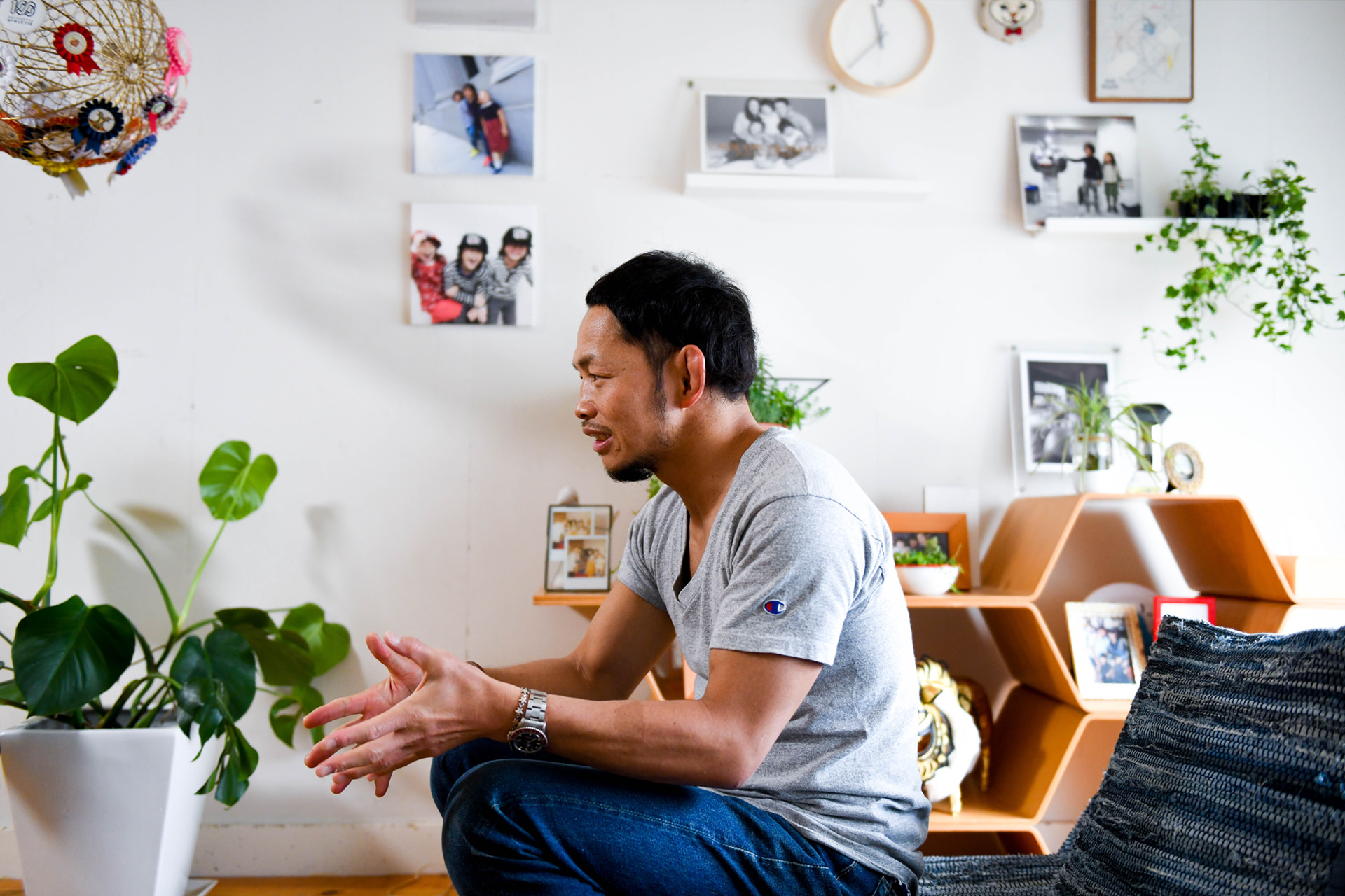 思い出の写真や切り抜きが壁一面にコラージュされた空間で、リラックスして座る男性の引きのショット。