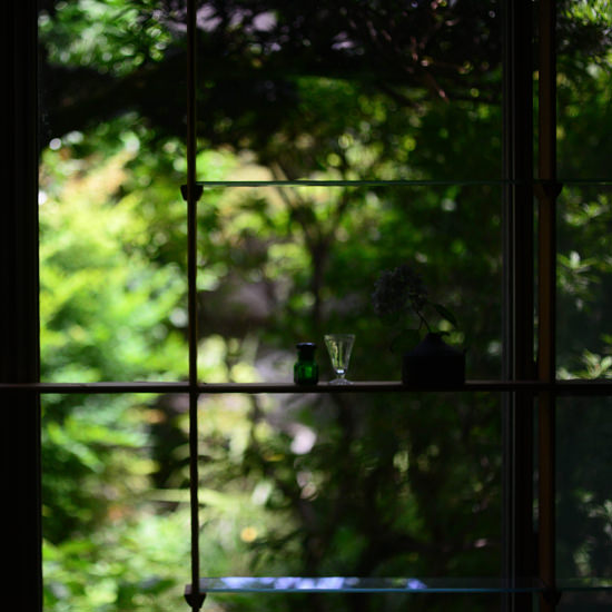 緑豊かな庭を背景に、窓際の棚に小さな瓶と一輪挿しが置かれた静かな風景。