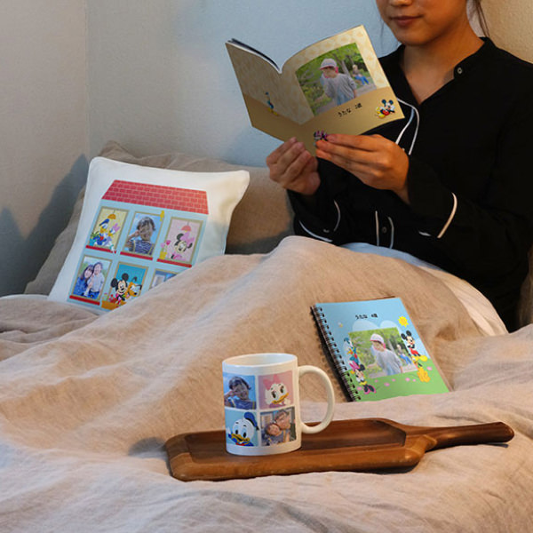 ベッドでフォトブックを広げる女性と、ディズニーデザインのマグカップやクッション