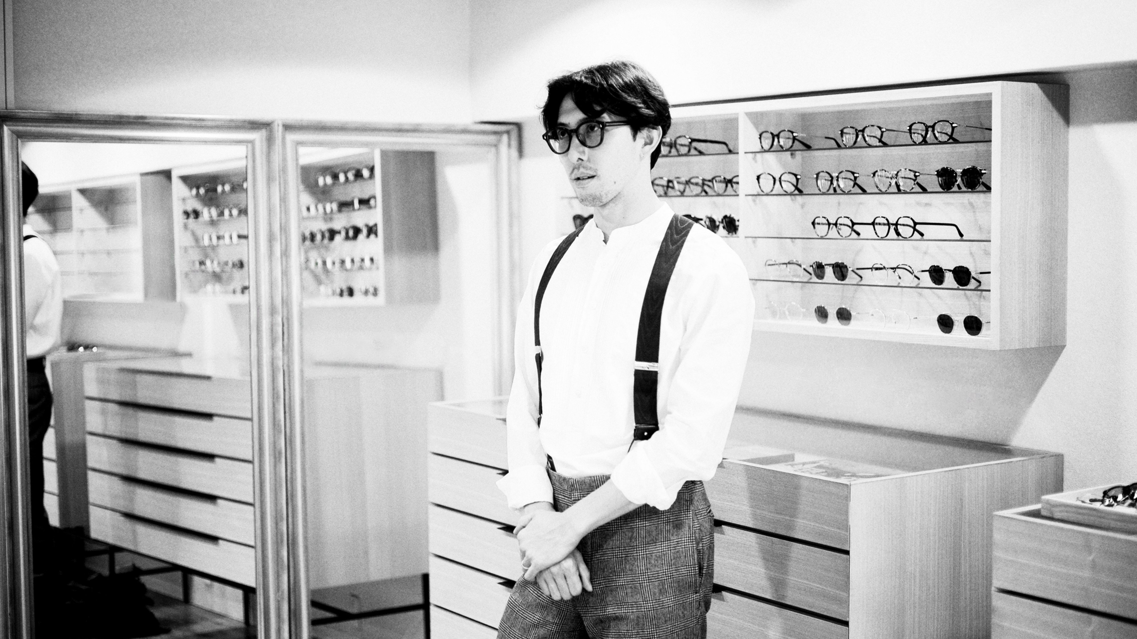 多数の眼鏡が並ぶ店内に立つ、ayameデザイナー今泉悠。背後の棚や鏡に整然とディスプレイされた眼鏡が写り込んでいる。