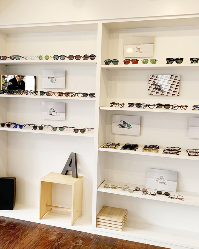 白い壁面の棚に多数の眼鏡やサングラスが整然と並び、その合間に眼鏡を主題としたアートパネルがいくつか飾られている。