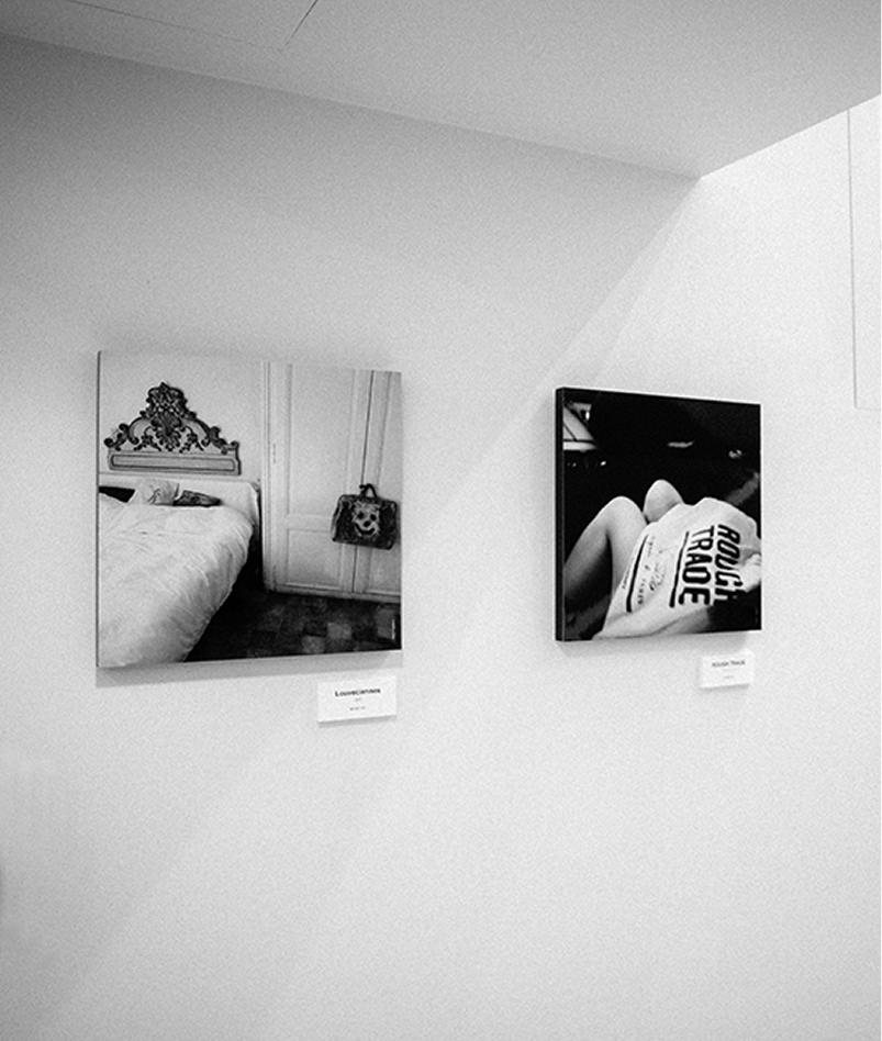 ギャラリーの白い壁面に展示された2枚のモノクロ写真パネル。左側は装飾的なヘッドボードのあるベッドルーム、右側は「ROUGH TRADE」の文字が入った服の裾などを写している。