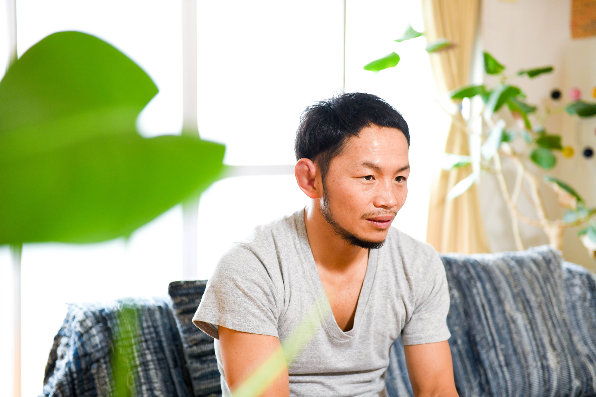 観葉植物の緑が映える明るい室内で、椅子に座りリラックスした表情を見せる男性。