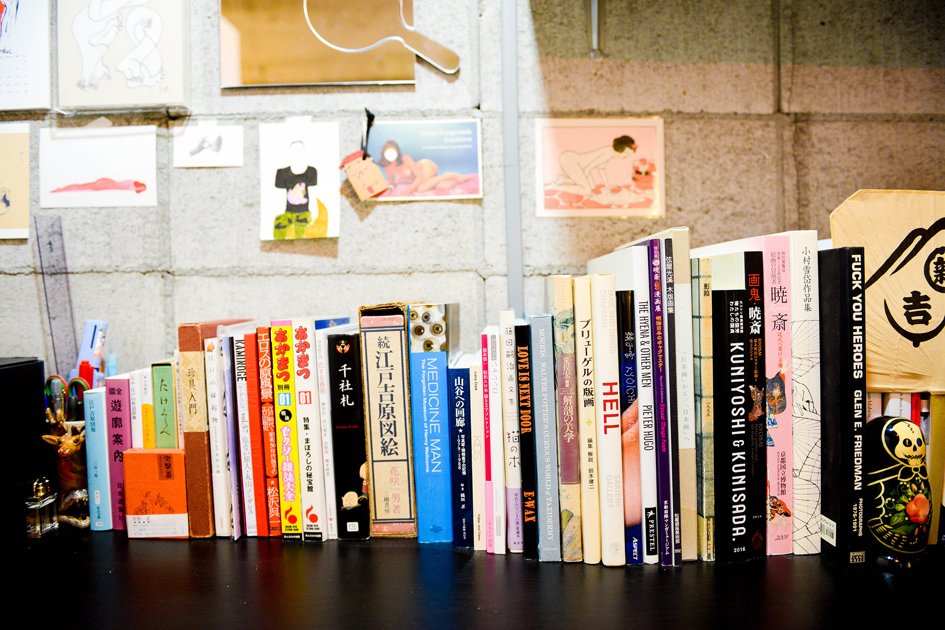 「続・江戸吉原図絵」や「KUNIYOSHI & KUNISADA」など、和洋の美術書や資料が並ぶ本棚。背後のコンクリート壁には様々なポストカードやイラストが飾られている。