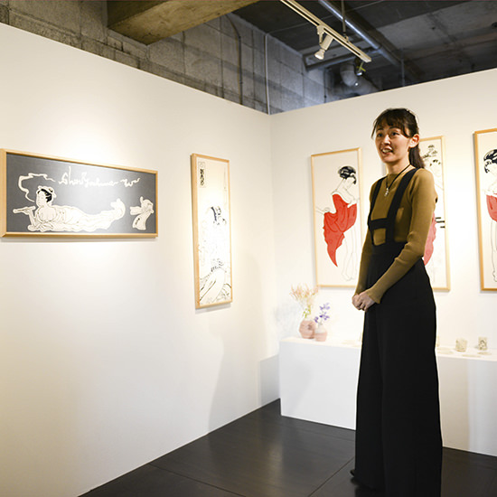 浮世絵風のイラストが描かれたアートボードが飾られた展示室に立つ女性。茶色のトップスに黒のサスペンダーパンツを着用した、洗練されたショップ内の風景。