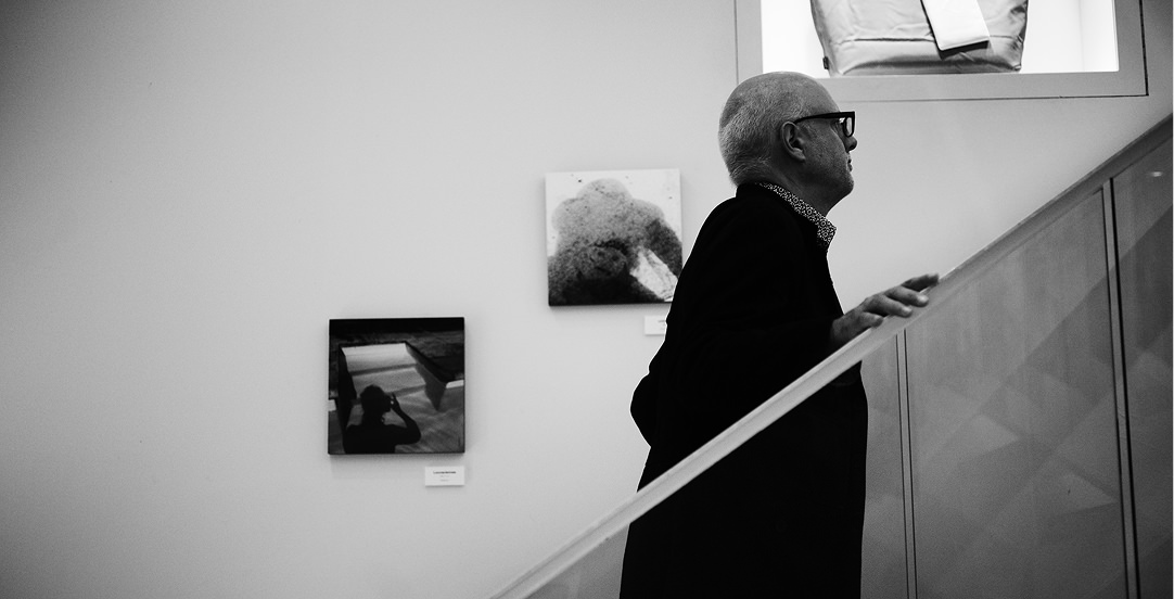 モノクロで表現された、眼鏡をかけた年配の男性が階段を上る横顔。背景の白い壁には、水面の反射や抽象的な質感を捉えた2枚の写真パネルが展示されている。
