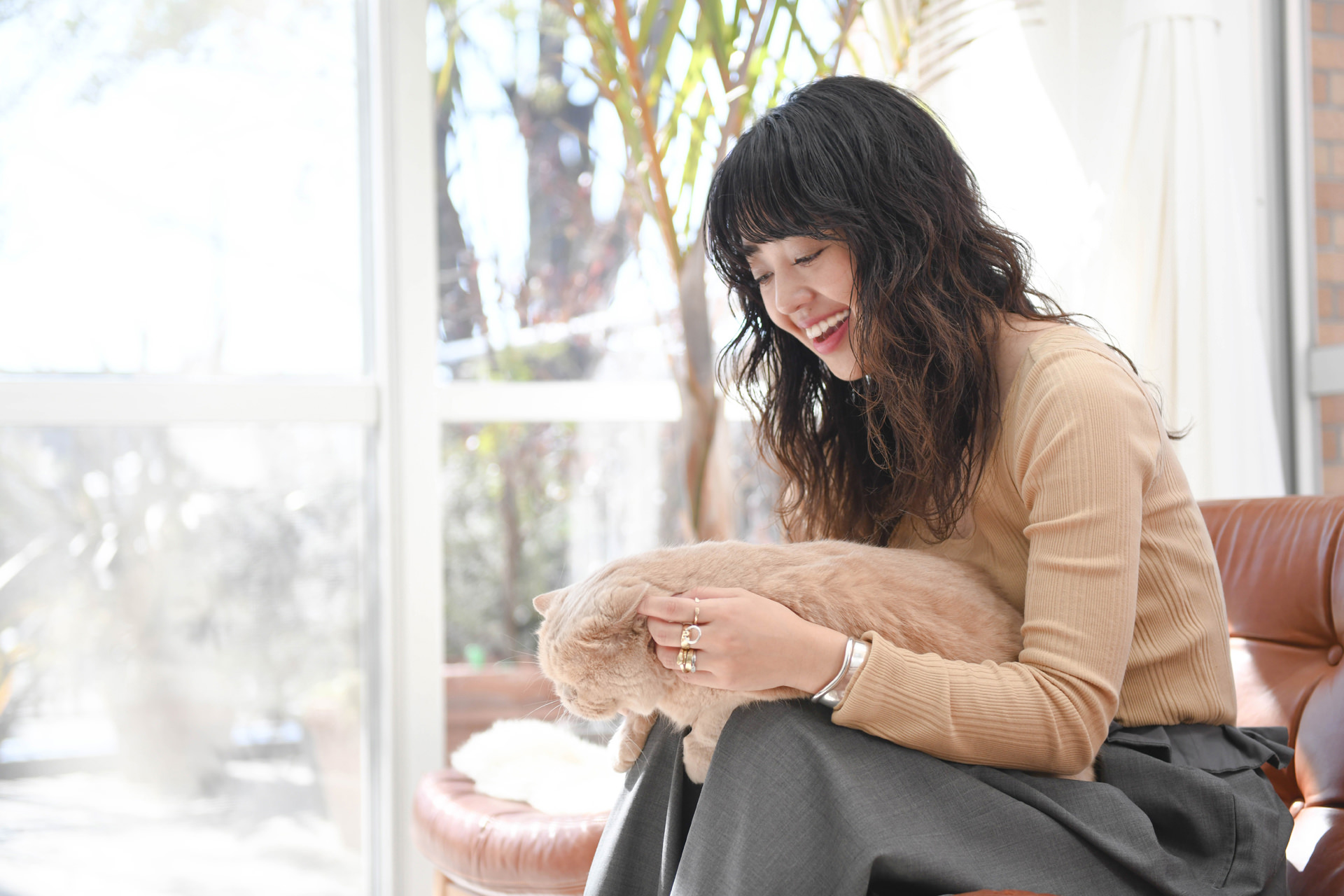 明るい窓際で、膝の上の猫を優しく撫でながら微笑む女性。穏やかな光が差し込む室内で、リラックスした表情を浮かべている。