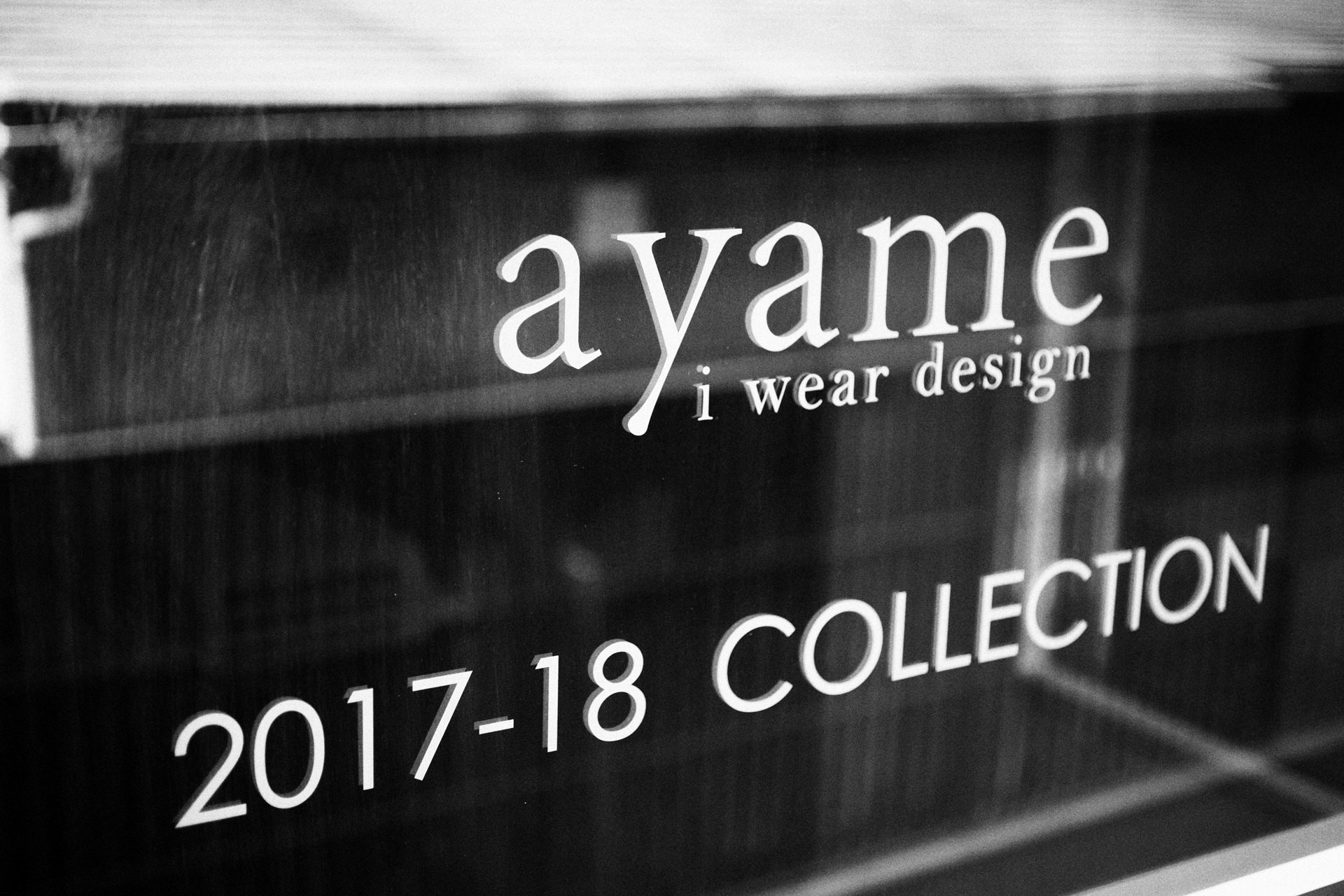 「ayame i wear design」および「17-18 COLLE...」のロゴが印字されたガラス面の接写。モノクロで表現された、スタイリッシュなショップサインの風景。
