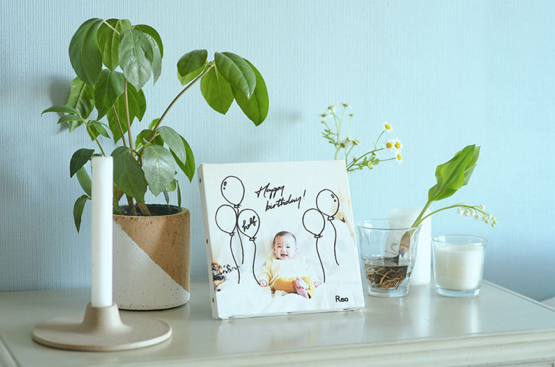 「Happy Birthday!」のメッセージと赤ちゃんの写真がプリントされたWALL DECOR キャンバス。水色の壁を背景に、観葉植物やキャンドルと一緒に飾られている。