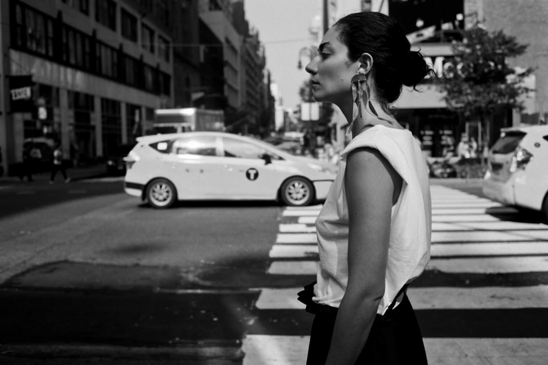 都会の横断歩道に立つ女性のモノクロポートレート。白いノースリーブに大きなイヤリングが印象的な横顔。背景にはタクシーが走る街並みが広がる。