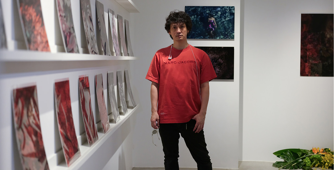 赤い「MARC JACOBS」のTシャツを着た安藤政信が、写真パネルが展示されたギャラリー内に立っている。背景の壁や左側の棚には、人物や花を主題とした写真作品が並んでいる。