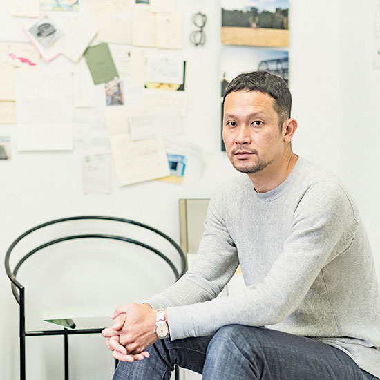 メモや写真、WALL DECOR ミュージアムが飾られたアトリエの壁を背に、手を組んで座る二俣公一。