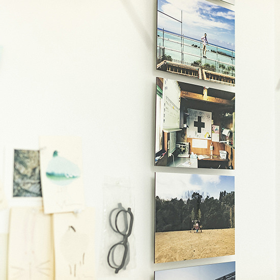 白い壁に縦一列に並んだWALL DECOR ミュージアムの接写。海辺や室内風景などを捉えた写真が並び、その傍らにはメモや眼鏡がディスプレイされている。