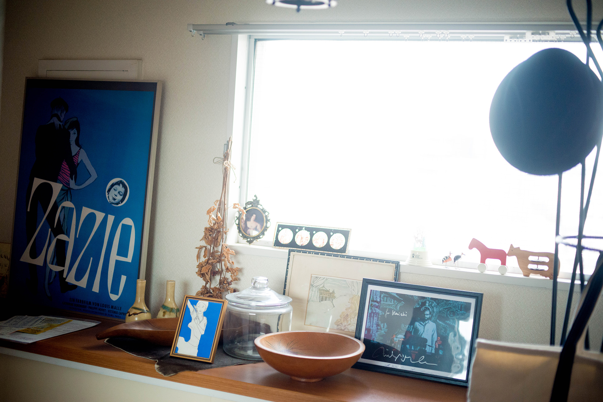 明るい窓際の棚に、青いポスターや額装写真、木製のボウルなどが並ぶインテリアの風景。