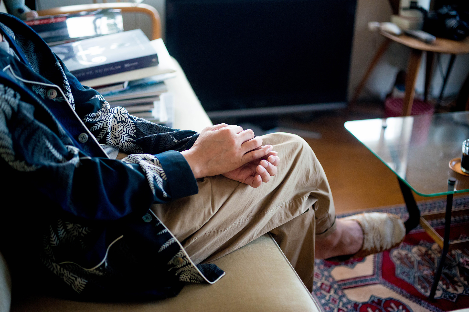 ソファーに座り、膝の上で静かに手を重ねる男性の手元のクローズアップ。足元に柄物のラグが広がる、リラックスした室内の風景。