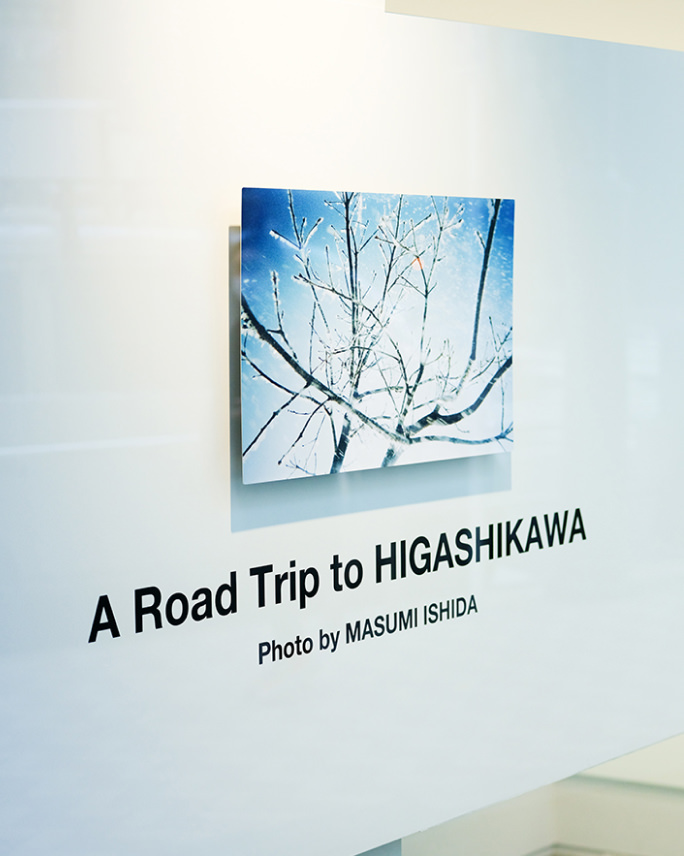 「A Road Trip to HIGASHIKAWA Photo by MASUMI ISHIDA」の文字が書かれた白い壁面に、冬の木々を写した青い色調の写真パネルが飾られている。