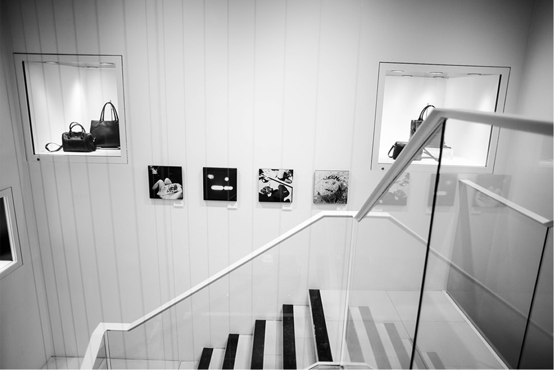 階段越しの壁面ディスプレイ。中央に4枚の小さな写真パネルが横一列に並び、その左右にある壁のくぼみ（ニッチ）には黒いハンドバッグが展示されている。