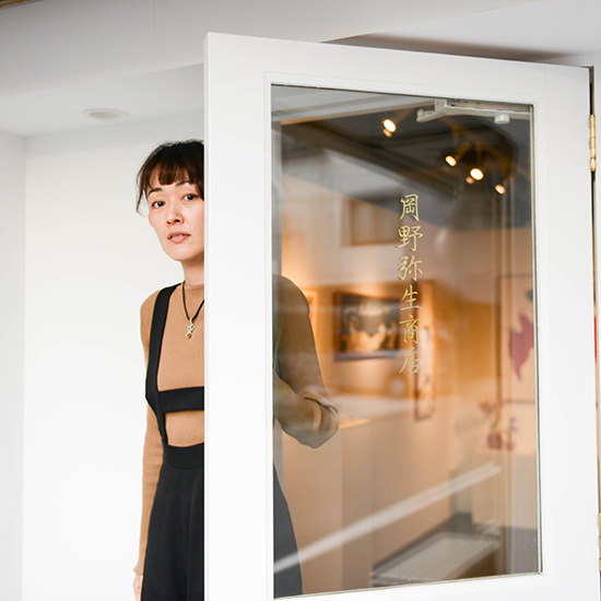 「月野静生商店」の金文字が書かれている白い扉から、静かに顔をのぞかせる女性のクローズアップ。