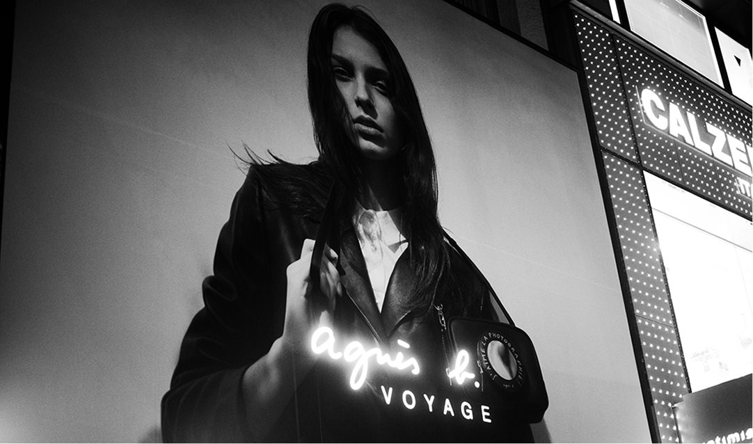 「agnès b. VOYAGE」のネオンサインが手前で白く光り、物憂げな表情の女性を捉えたモノクロの写真パネル。