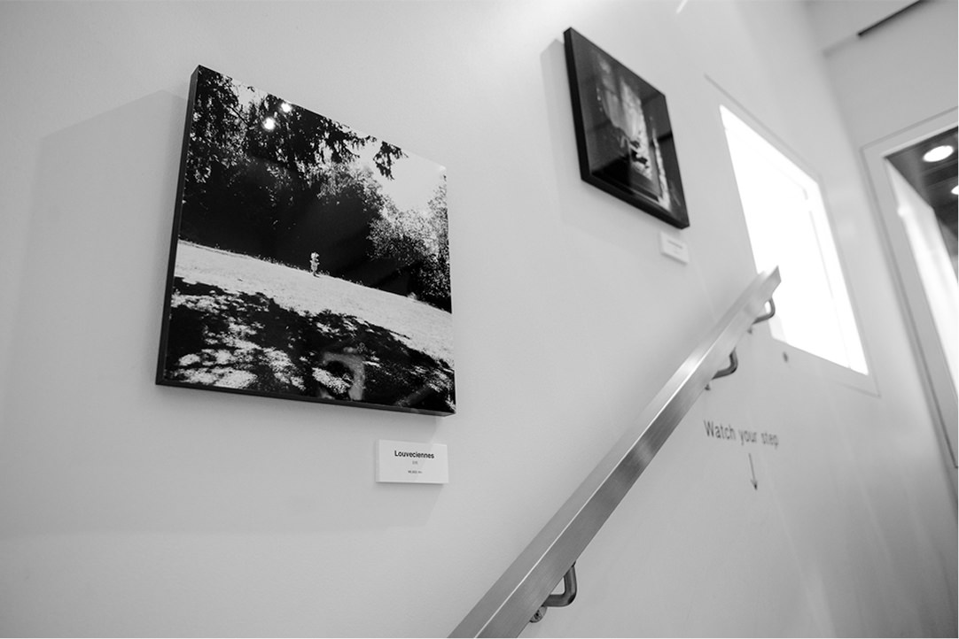 階段横の白い壁面に展示された2枚の写真パネル。左下の写真には広々とした庭園のような風景が写り、その下には「Louveciennes」と記された小さなプレートがある。壁面には「Watch your step」という注意書きと矢印が添えられている。