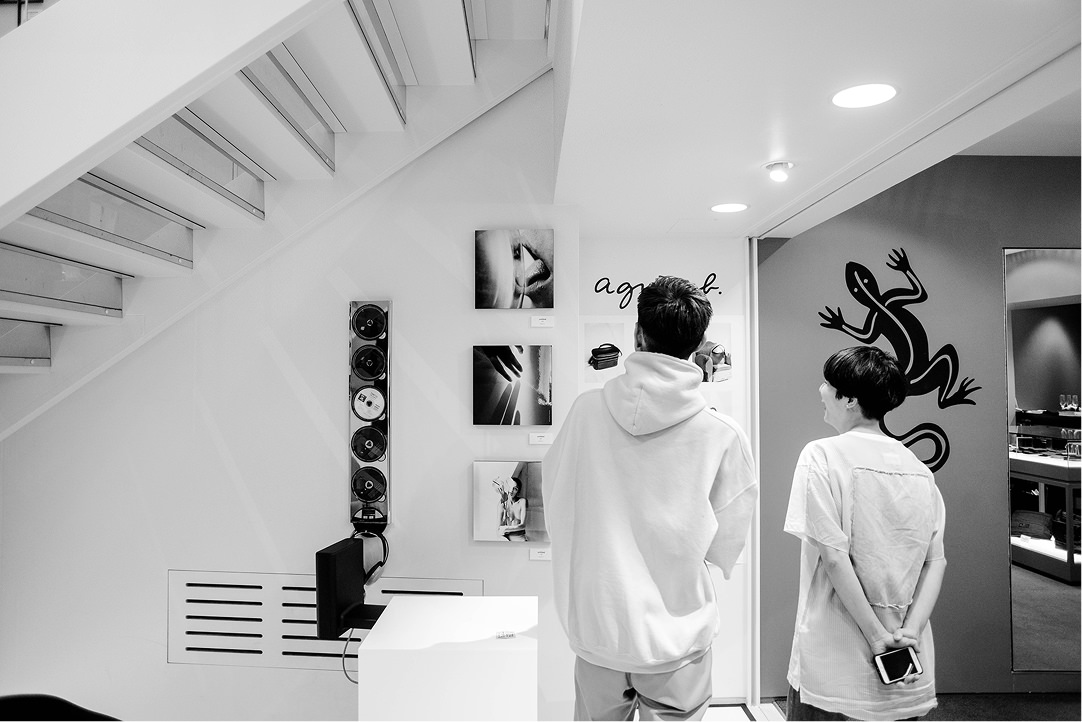 モノクロで表現された、階段下の展示スペース。白い壁面に3枚の写真パネルが縦に並び、その横には「agnès b.」のロゴや大きなトカゲのイラストが描かれている。手前では2人の人物が展示を鑑賞している。
