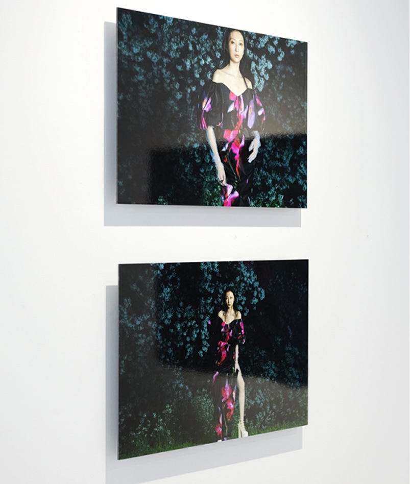 白い壁に縦に2枚並べて展示された、花々に囲まれて立つ女性の姿を写した写真パネル。