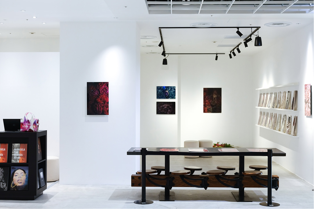 白い壁面に赤い花や人物の写真パネルが展示されたギャラリー空間。手前にはスツールが備え付けられた長いテーブルが置かれ、右側の棚にはパンフレットが並んでいる。