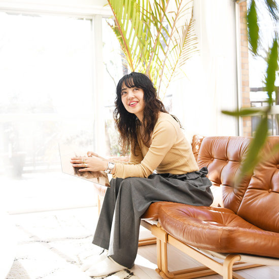 明るい日差しが差し込むリビングで、レザーソファに座り微笑む女性。手元には写真パネルを持ち、リラックスした表情。