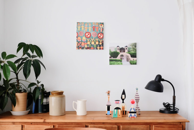 白い壁に2枚飾られた、カラフルなイラストと建物の写真のWALL DECOR キャンバス。観葉植物や小物が並ぶ棚の上にディスプレイされている。