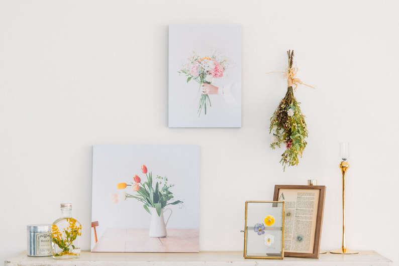 白い壁に2枚飾られた、花束や生け花の写真のWALL DECOR カジュアル。ドライフラワーのスワッグや小物が並ぶ空間にディスプレイされている。
