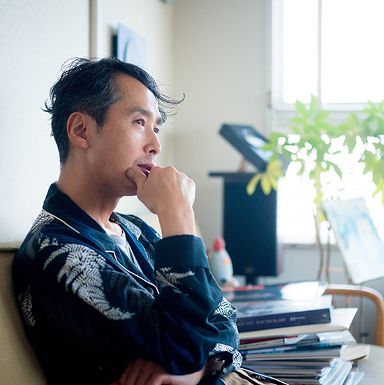 ソファーで顎に手を添え、静かに物思いにふける青野 賢一。背後の壁には円形のアートが飾られ、明るい窓際を背景にした室内の風景。