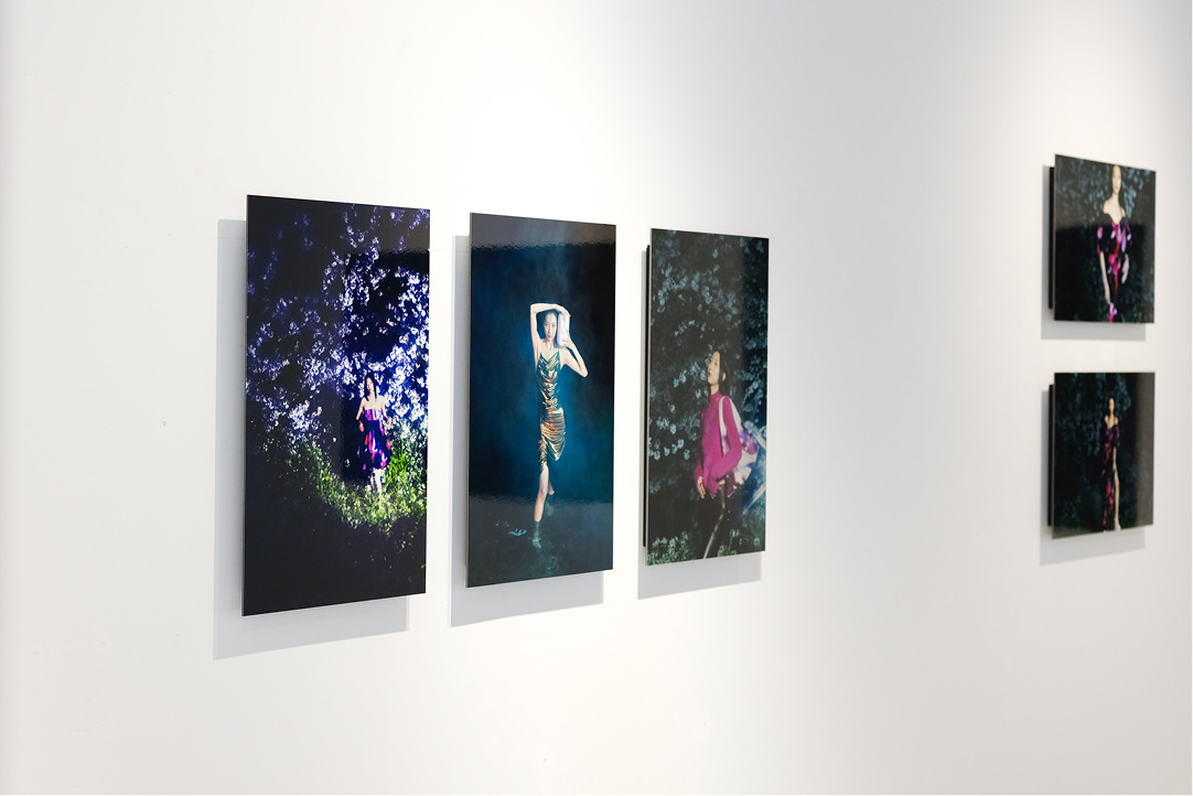 ギャラリーの白い壁面に、煌びやかなドレスやピンクの服を纏った女性を写した3枚の縦長写真パネルが横に並んで展示されている。
