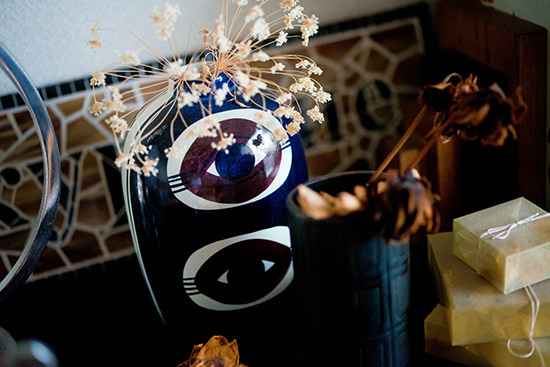 目の模様が描かれた青い花瓶にドライフラワーが活けられ、黒い器や包まれた小物が並ぶ芸術的な卓上のクローズアップ。