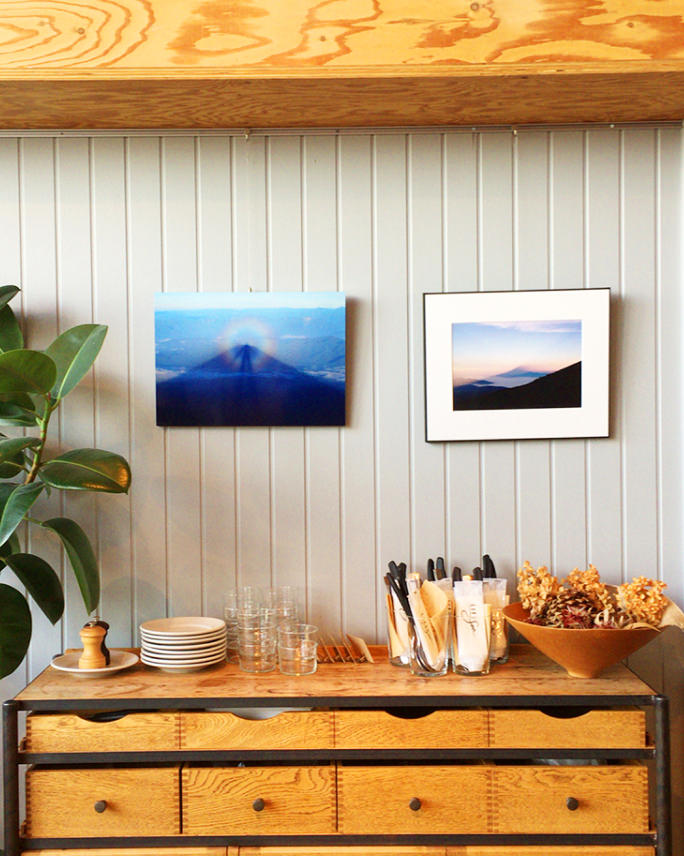 白い板壁に山の風景写真のパネルを2枚飾り、下の木の棚には食器やカトラリー、植物などが整然と並んでいる。