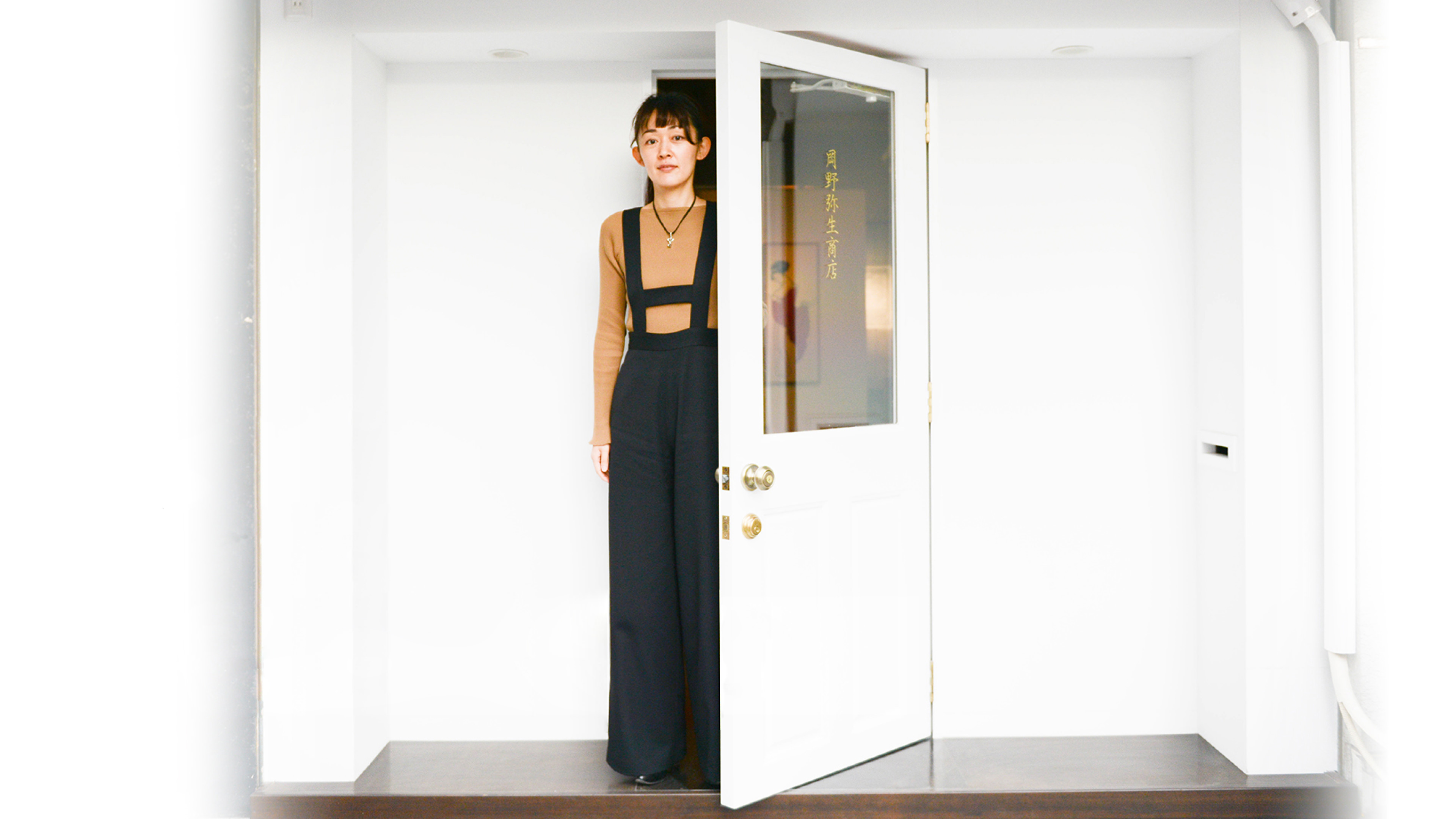 「月野静生商店」と金色の文字が記された白い扉の入り口に立つ岡野弥生。茶色のトップスと黒のサスペンダーパンツを身にまとったポートレート。