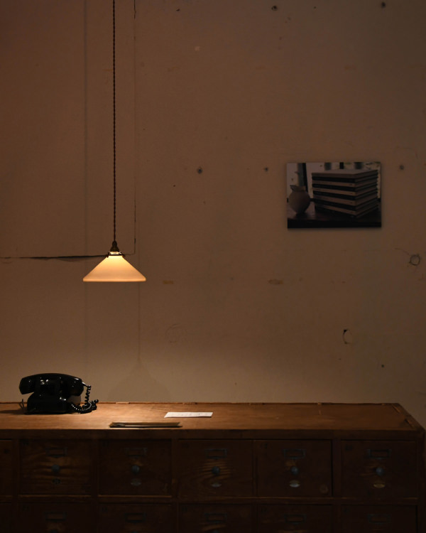 ペンダントライトが照らす薄暗い部屋で、壁に本の写真のWALL DECORを飾り、棚に黒い電話機を置いている。