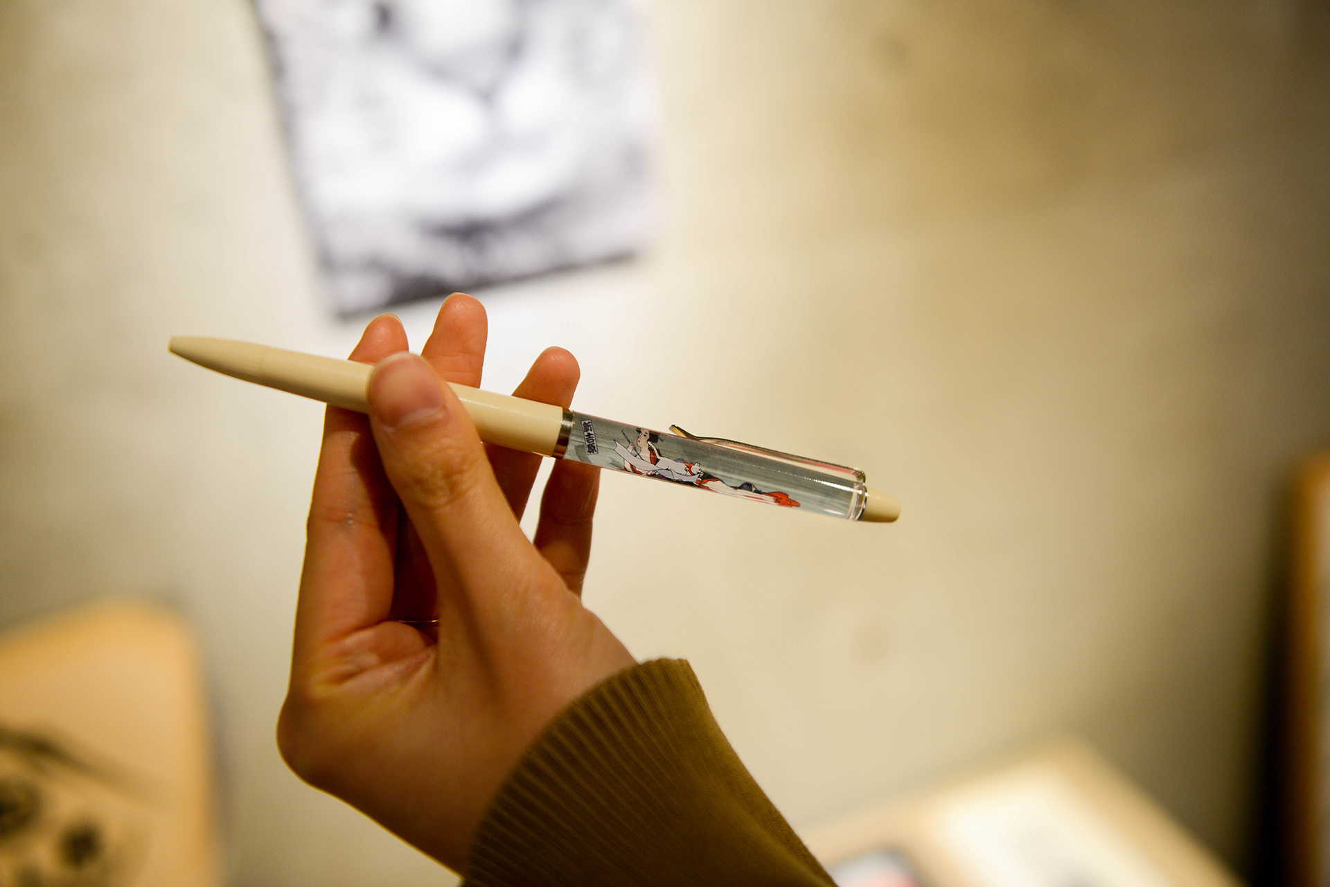 伝統的な浮世絵風の女性が描かれた、透明なパーツ付きの白いボールペンを持つ手。背景にはコンクリート壁がぼけて写っている。