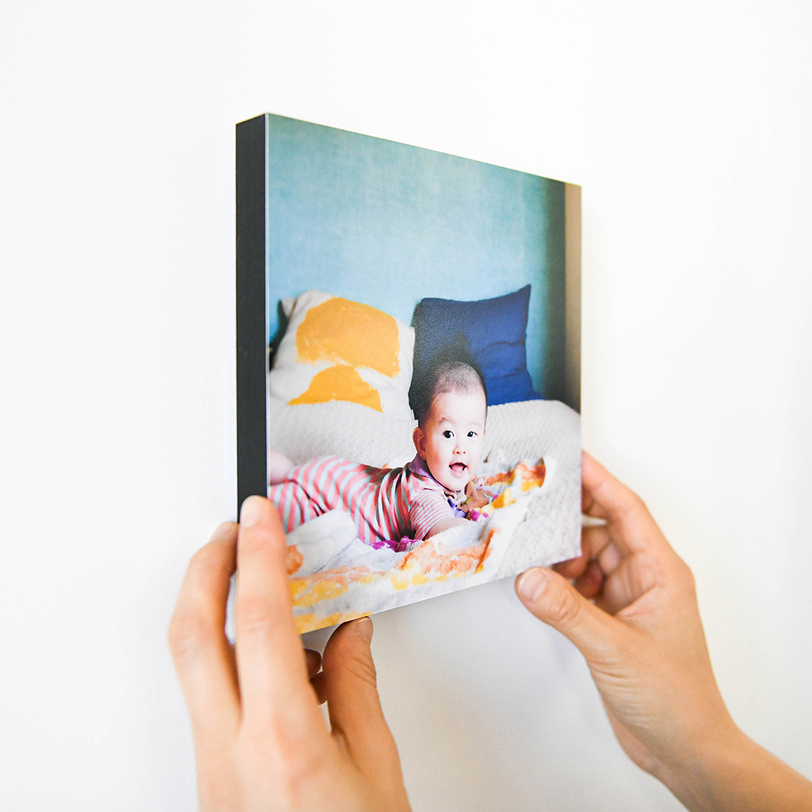 ベッドでうつ伏せになった赤ちゃんの写真のWALL DECOR カジュアルを、壁に飾ろうと両手で持っている様子