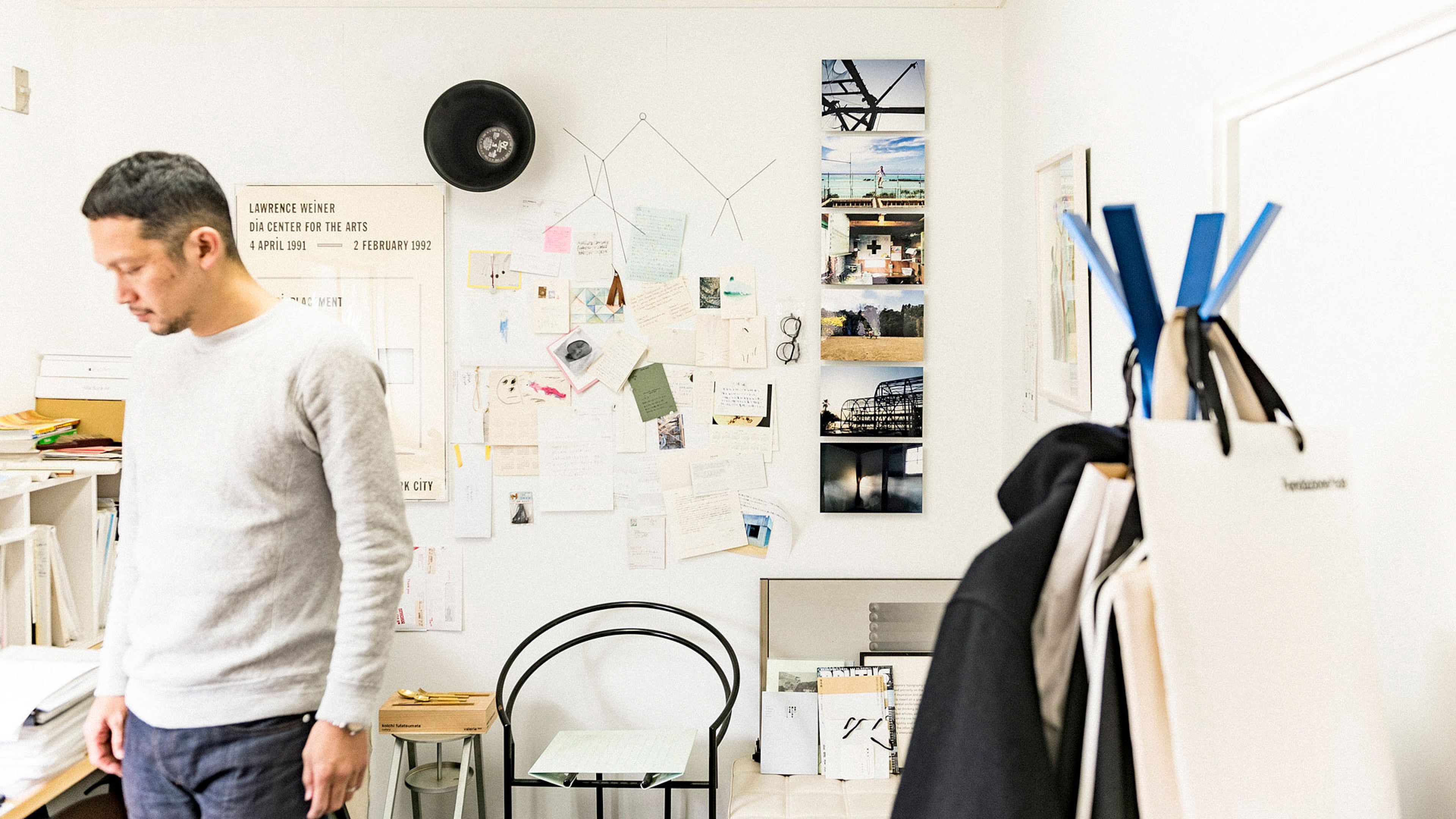 壁一面に写真やメモ、スケッチが飾られたクリエイティブなアトリエの風景。手前に立つ男性と、右側に置かれた青いアクセントのコートハンガーが写り込んでいる。