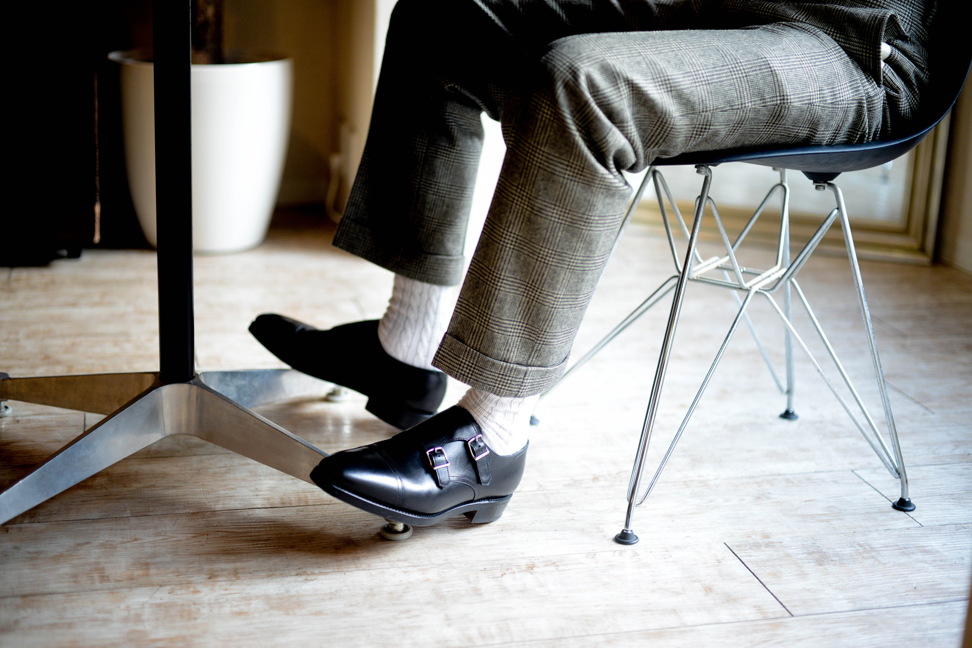 椅子に座る男性の足元のクローズアップ。チェック柄のパンツに白い靴下、黒い革靴を履き、デザイン性の高いワイヤー脚の椅子に腰掛けている。
