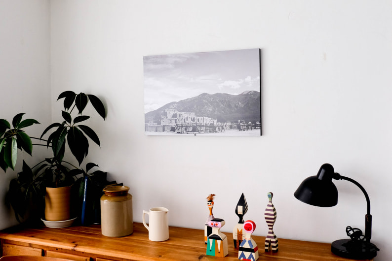 白い壁に飾られた、山と建物のモノクロ写真のWALL DECOR カジュアル。観葉植物や小物が並ぶ棚の上にディスプレイされている。