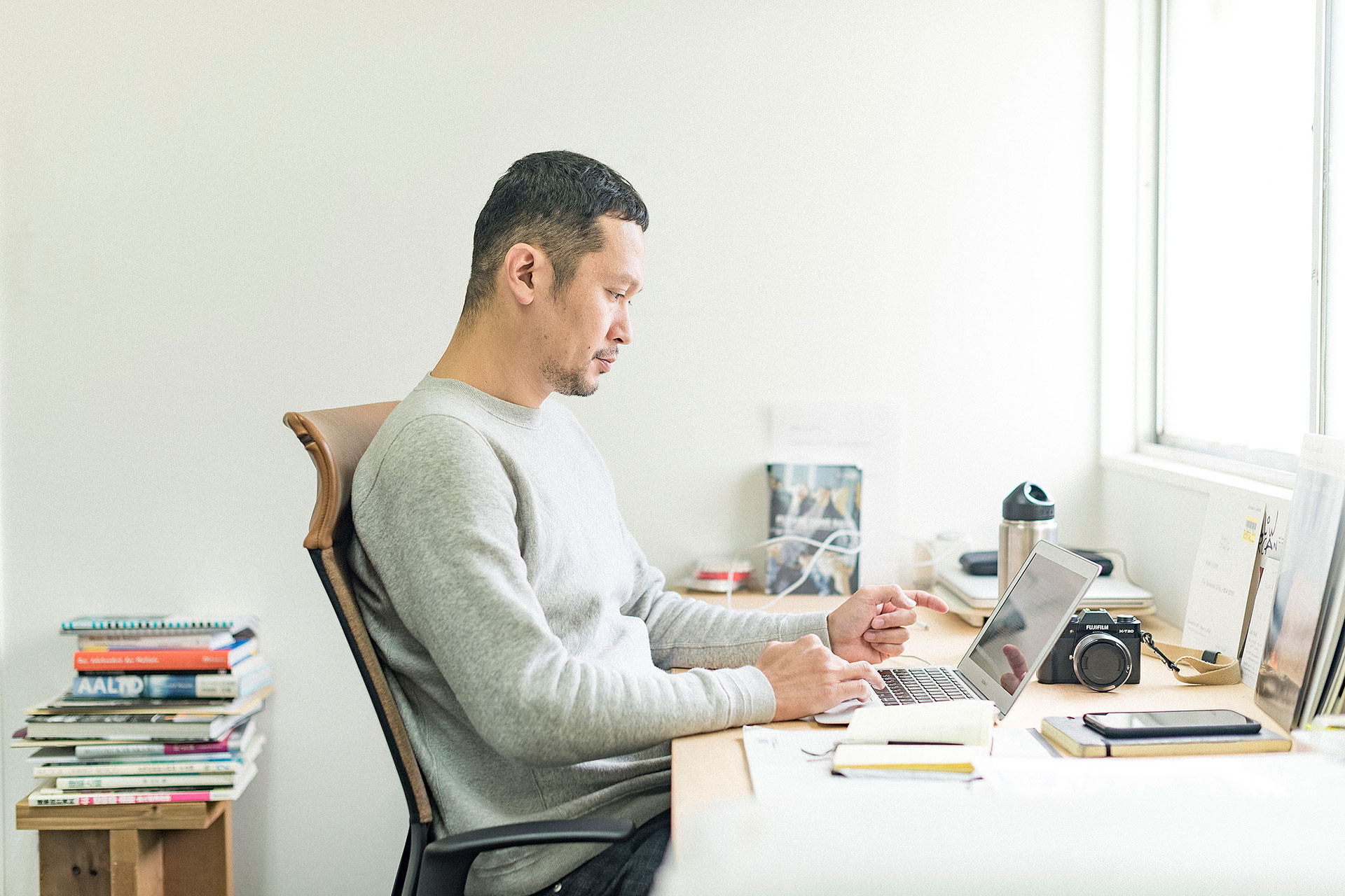 明るい窓際のデスクでノートパソコンを操作し、作業に集中する二俣公一。手元にはカメラやスマートフォンがあり、傍らには本が積み上げられた仕事場の風景。