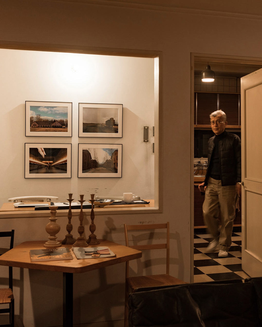手前に木のテーブルと燭台があり、壁に4枚の風景写真のWALL DECORを飾った部屋を歩く男性。