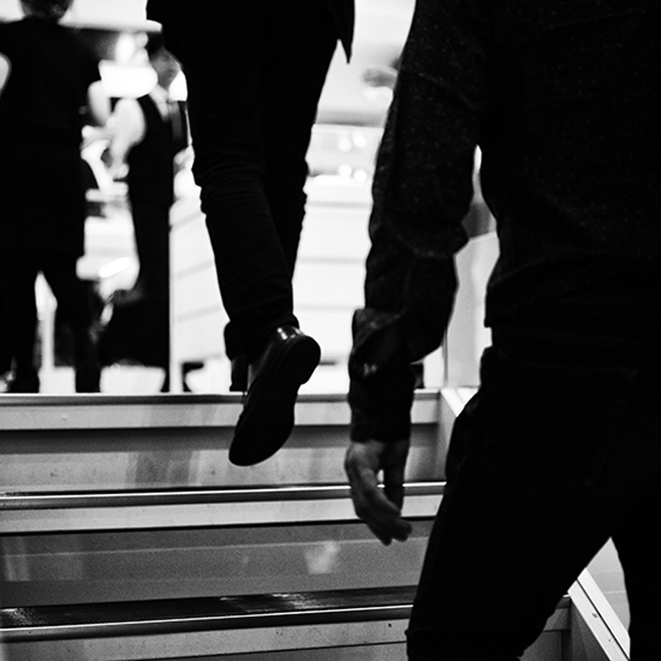 モノクロで表現された、階段を上る人々の足元と下半身のクローズアップ。片方の足が次の一段へ踏み出される瞬間を捉えている。