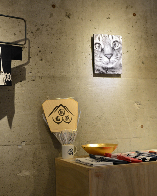 コンクリートの壁に猫の顔のモノクロ写真のWALL DECORを飾り、手前の木の棚にうちわや小物を並べる。