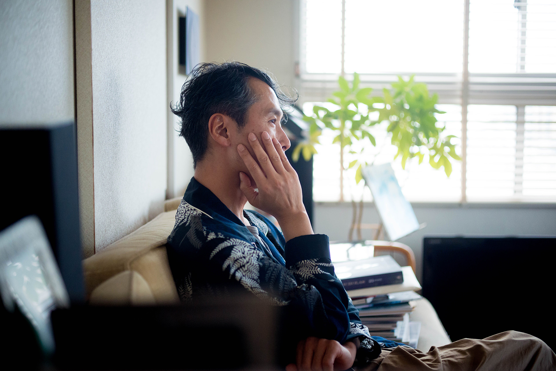 ソファーで頬杖をつき、静かに窓の外を見つめる男性。自然光が差し込む明るい室内に、観葉植物や積み上げられた本が置かれている。