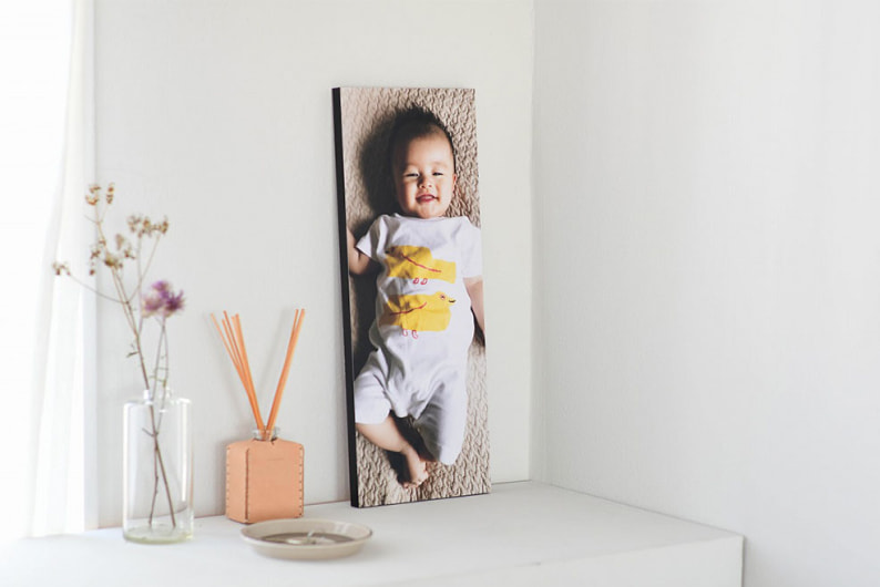 白い棚に飾られた、笑顔の赤ちゃんの写真の縦長なWALL DECOR カジュアル。ディフューザーやドライフラワーが添えられている。