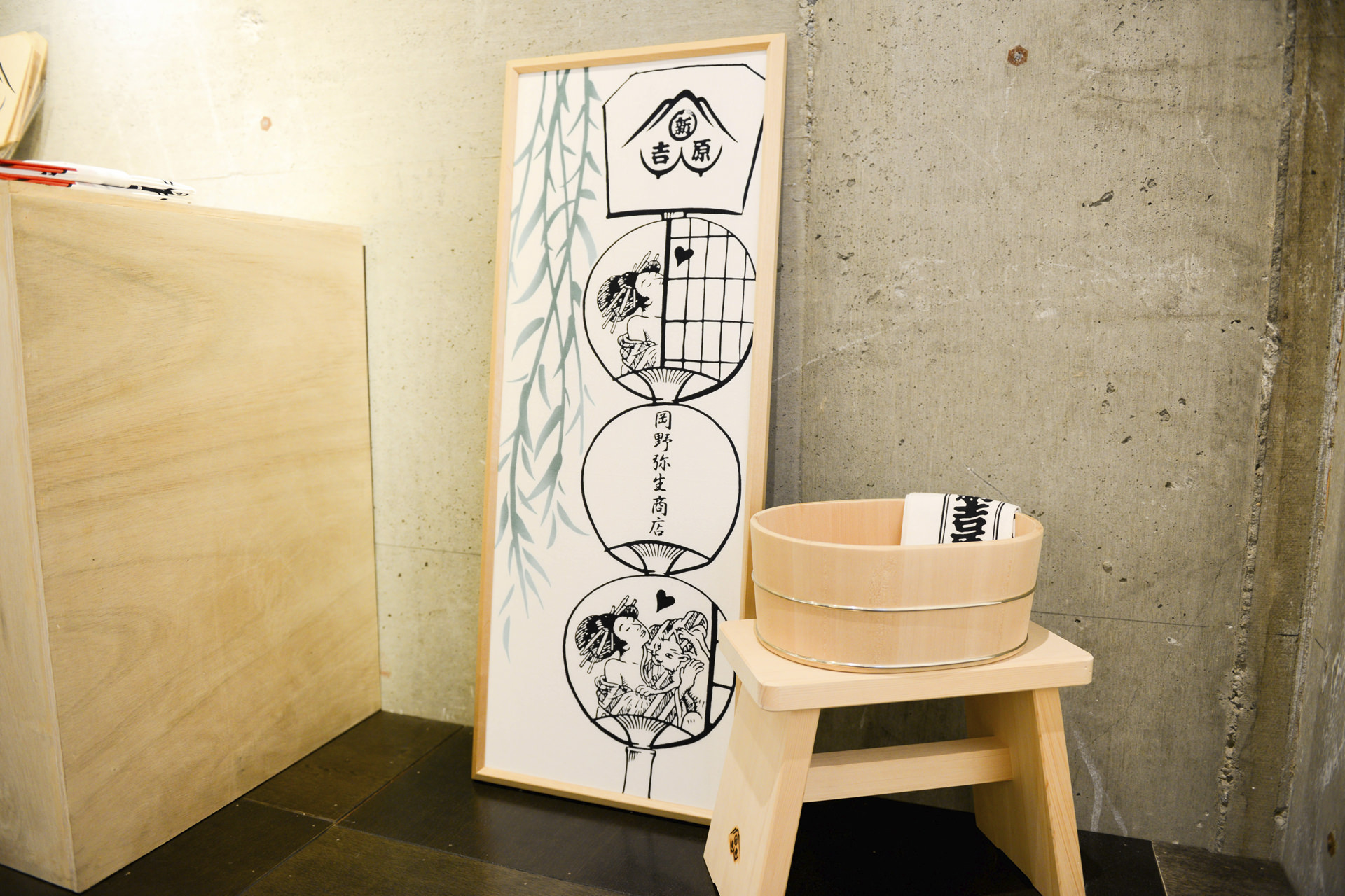 「新吉原」と「月野静生商店」の文字、そして団扇の中に描かれた女性のイラストが特徴的な縦長のアートボード。その手前に木製の湯桶と風呂椅子が置かれた、和の情緒漂うディスプレイ風景。
