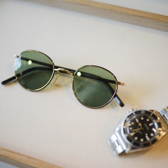 トレイに置かれた、細いゴールドと黒のフレームにグリーンのレンズを組み合わせた丸型サングラス。傍らにシルバーの腕時計が置かれている。