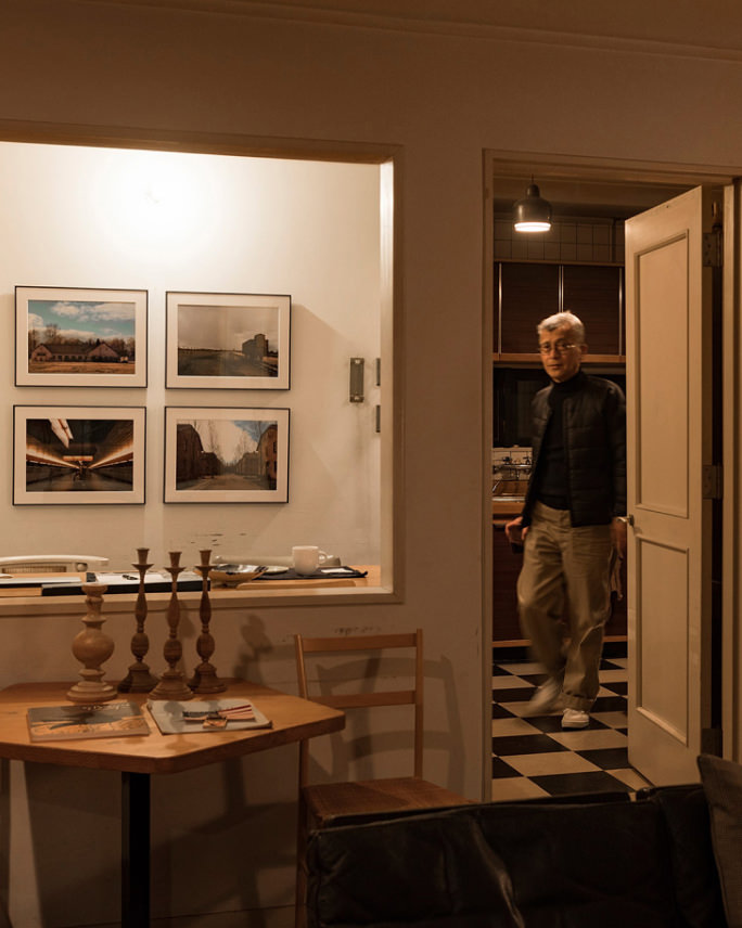 手前に木のテーブルと燭台があり、壁に4枚の風景写真のWALL DECOR グロッシーを飾った部屋を歩く男性。